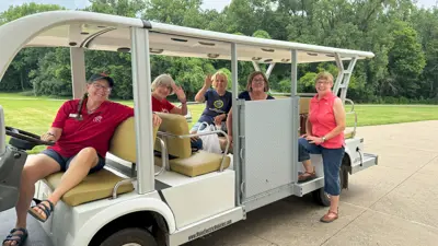 Group of five people seated and standing around a white open-air tram parked on a paved surface, with grassy fields and trees in the background. Several of the individuals are seated inside the tram while one stands nearby, and the vehicle is positioned outdoors under daylight.
