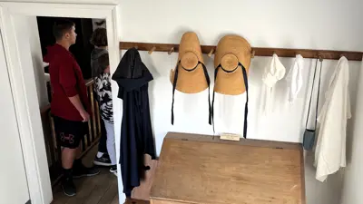 Historic interior room with wooden plank floors and white walls featuring a simple wooden bench and desk. A wall-mounted rack holds straw hats and garments, while two visitors stand in a doorway at the back of the room looking into another space.