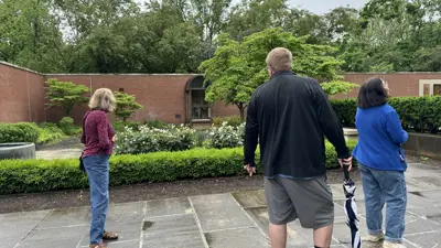 Three visitors stand on a wet paved courtyard, looking toward a circular garden feature bordered by low hedges in a walled outdoor space. One visitor in the foreground holds a folded umbrella, while a brick building and lush trees frame the background under an overcast sky.