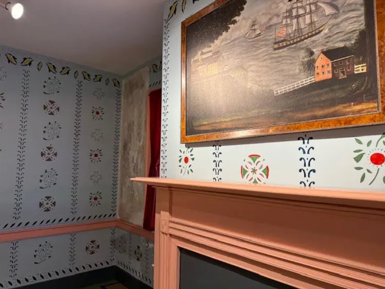 Interior of a historic room with a painted mantel and decorative stencil patterns on pale walls, featuring a framed painting of a river scene with boats and a building, and a window with red curtains in the background.