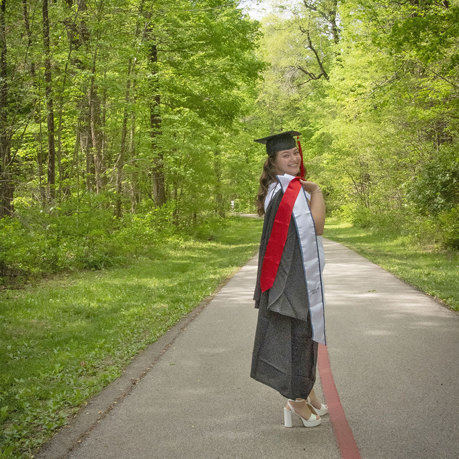 A 2025 graduate looks over her shoulder, her gown and honors stole thrown over her shoulder on the paved USI/Burdette Trail 