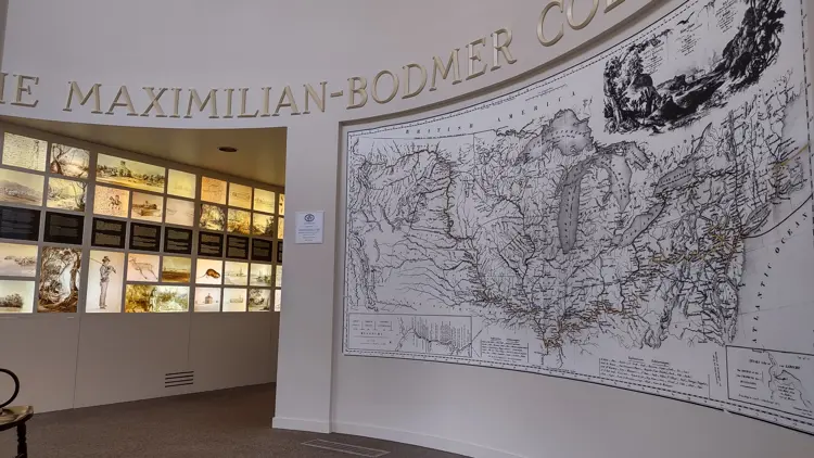 Interior of the Atheneum exhibit space featuring a large wall-mounted historical map and the text “Maximilian-Bodmer Collection” above, with illuminated panels displaying artwork and illustrations along a curved gallery wall.