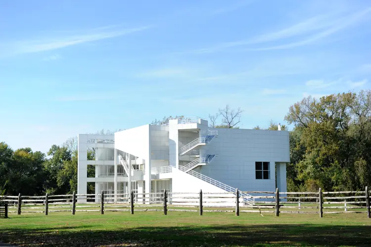 The white modernist Atheneum building stands behind a wooden fence and open lawn, with exterior staircases and geometric forms visible against a clear blue sky.