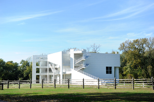 A white, modernist building with clean, geometric lines and an exterior staircase stands behind a wooden fence in a grassy landscape. Large windows wrap around one corner of the structure, and trees with full foliage surround the building under a clear blue sky.