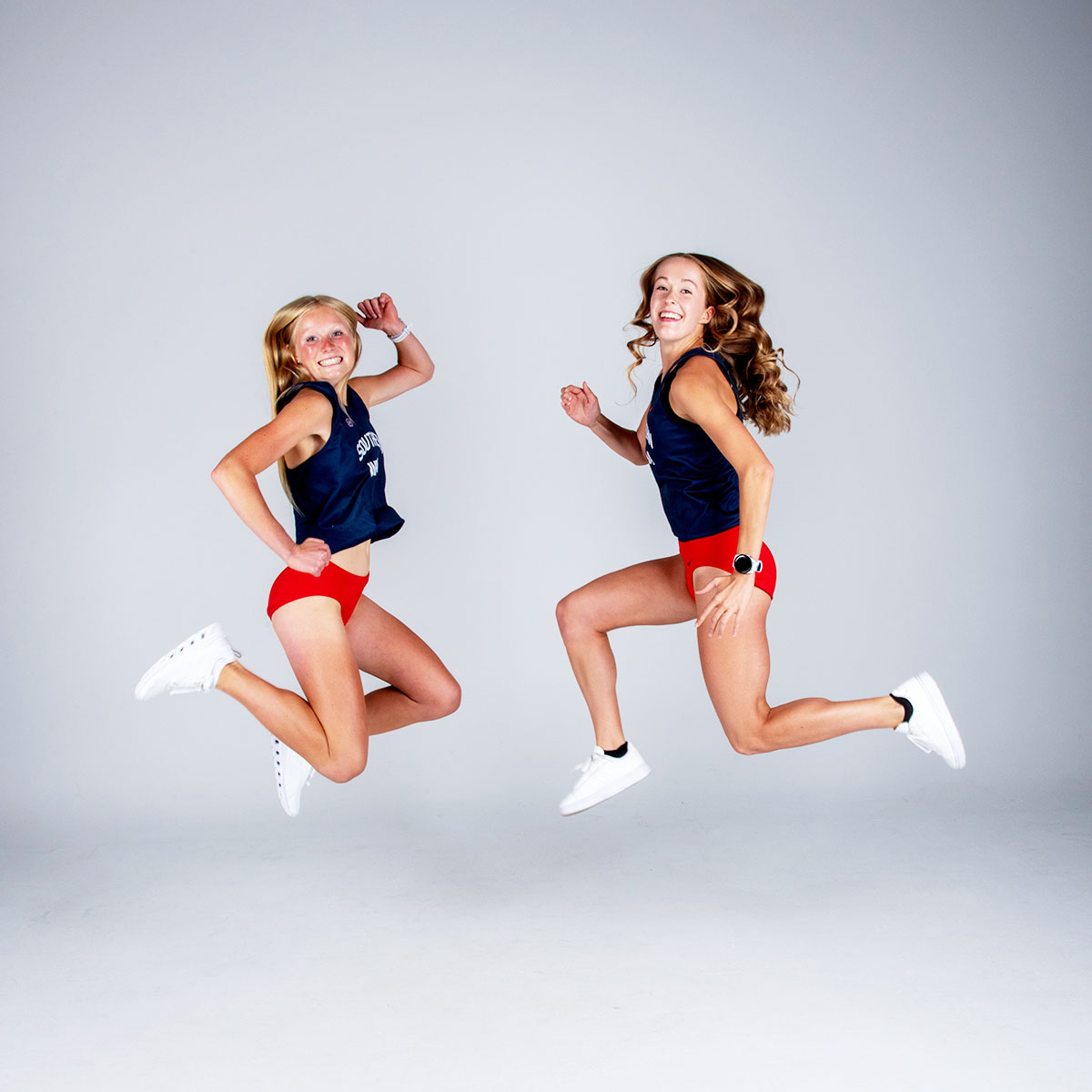 Two female cross country runners jump high off the ground for their media photos. 