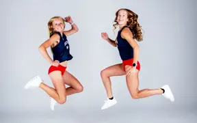 Two female cross country runners jump high off the ground for their media photos. 