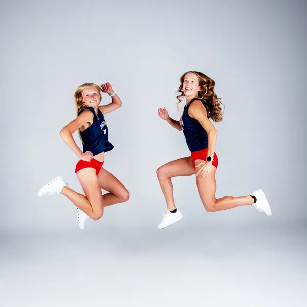 Two female cross country runners jump high off the ground for their media photos. 