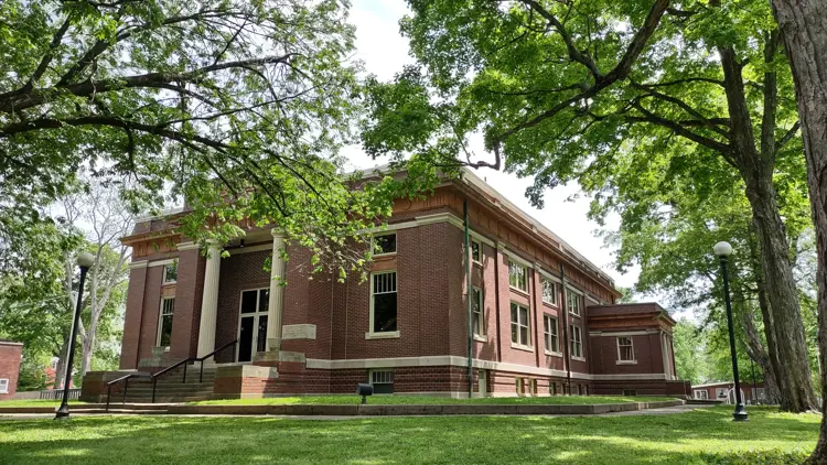 A historic red brick building with white columns and symmetrical windows stands beneath leafy trees, with a front entrance reached by steps and a lawn and walkway in the foreground.