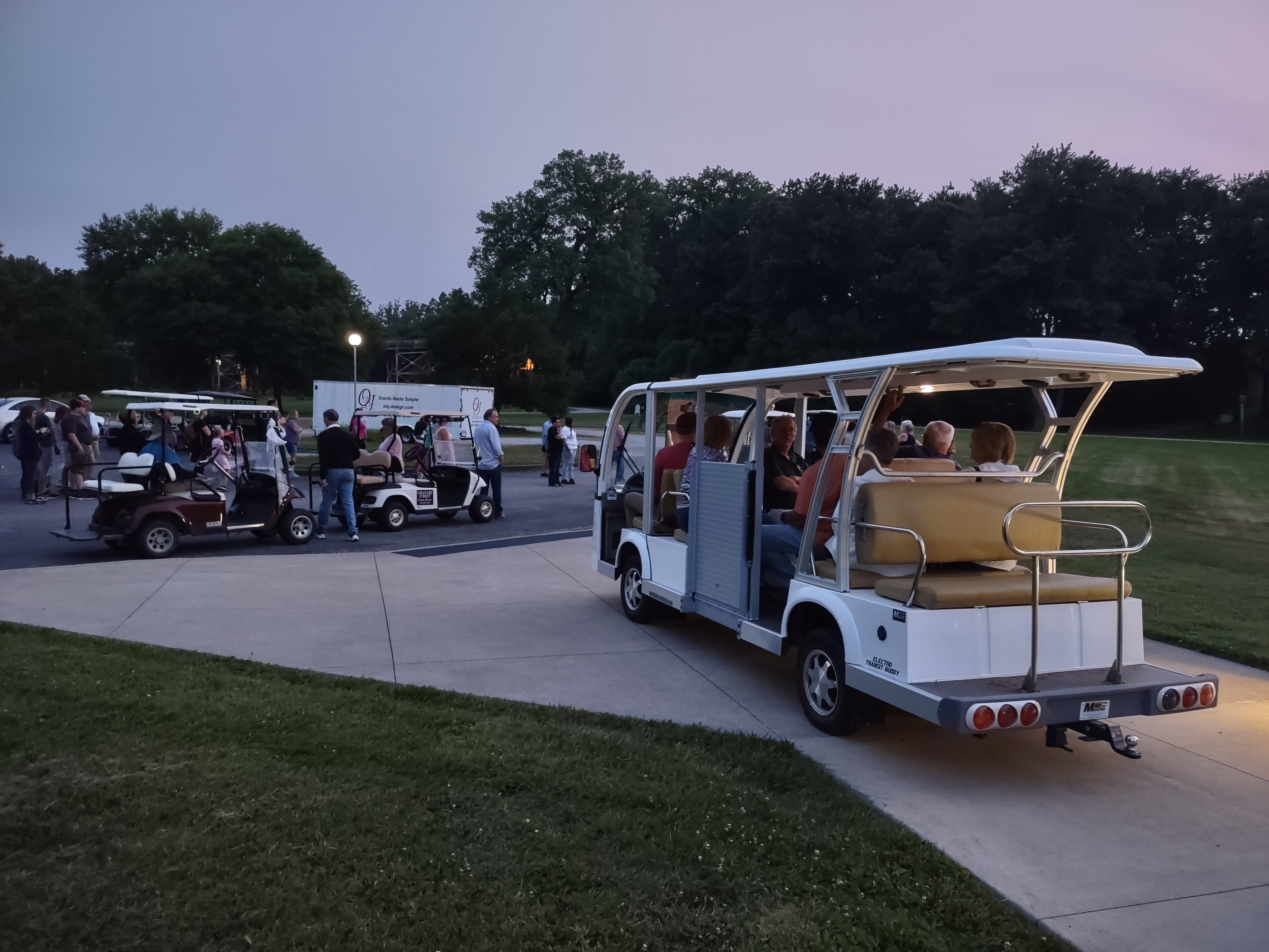 Golf carts and a tram transporting visitors along a paved path at dusk with people nearby.