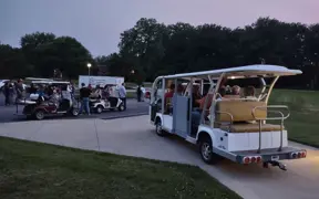 Golf carts and a tram transporting visitors along a paved path at dusk with people nearby.