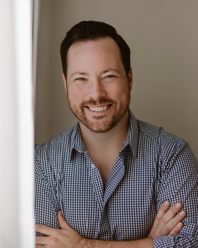 Craig Fehrman head shot; white man, dark hair and facial hair, wearing a gingham button down shirt