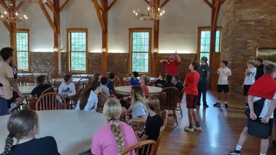 A large group of visitors sits and stands inside a spacious room with exposed wooden beams and tall windows, gathered around white round tables and wooden chairs. At the front of the room, a guide gestures while speaking to the group. Chandeliers hang from the ceiling, and the warm wood flooring and brick walls create a historic, lodge-like setting.