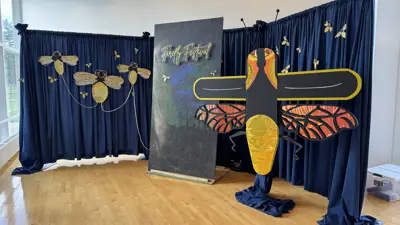 Indoor festival display featuring a large stylized firefly sculpture with black wings and orange detailing, set in front of dark blue curtains decorated with gold firefly shapes. A vertical sign reading “Firefly Festival” stands behind the sculpture, and natural light enters from tall windows on the left, illuminating the wooden floor.