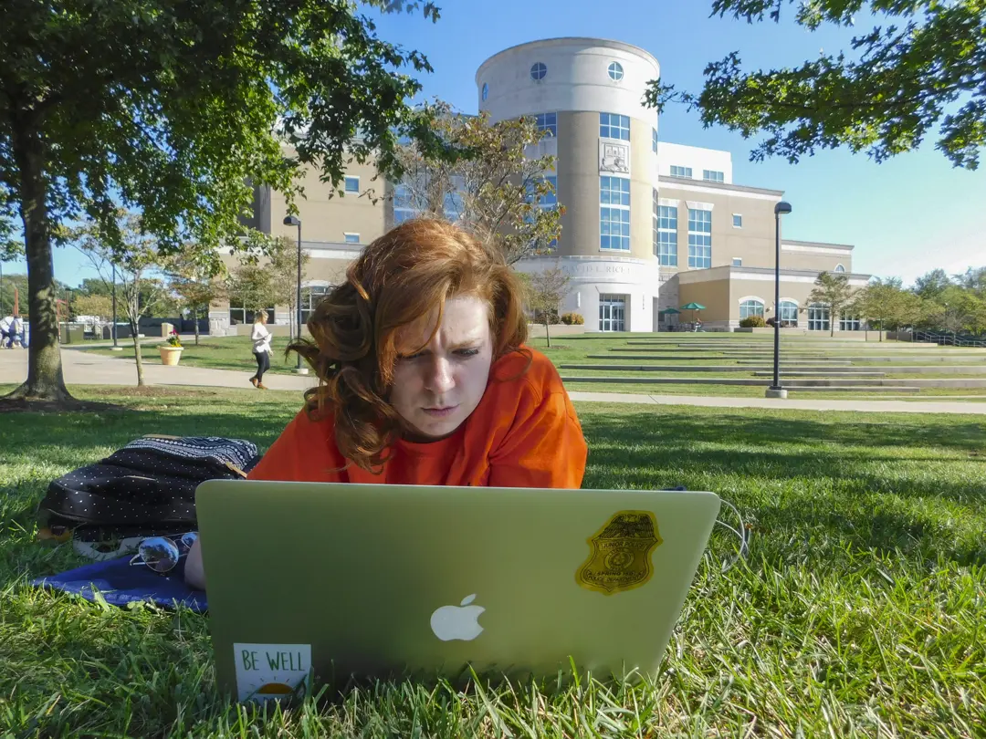 Student laying on the grass on the Quad working on her laptop with Rice Library in the background