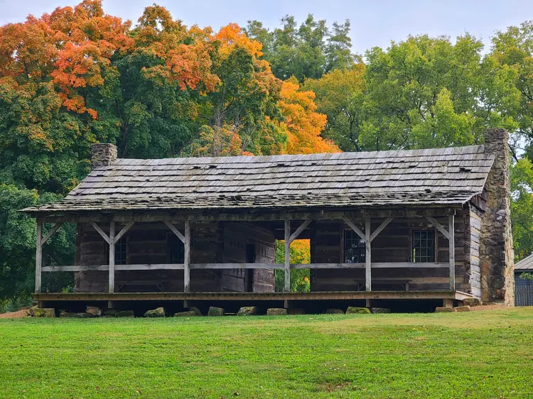 A long, weathered log building with a covered front porch and stone chimneys stands on a grassy lawn, with colorful autumn trees in shades of green and orange rising behind it.