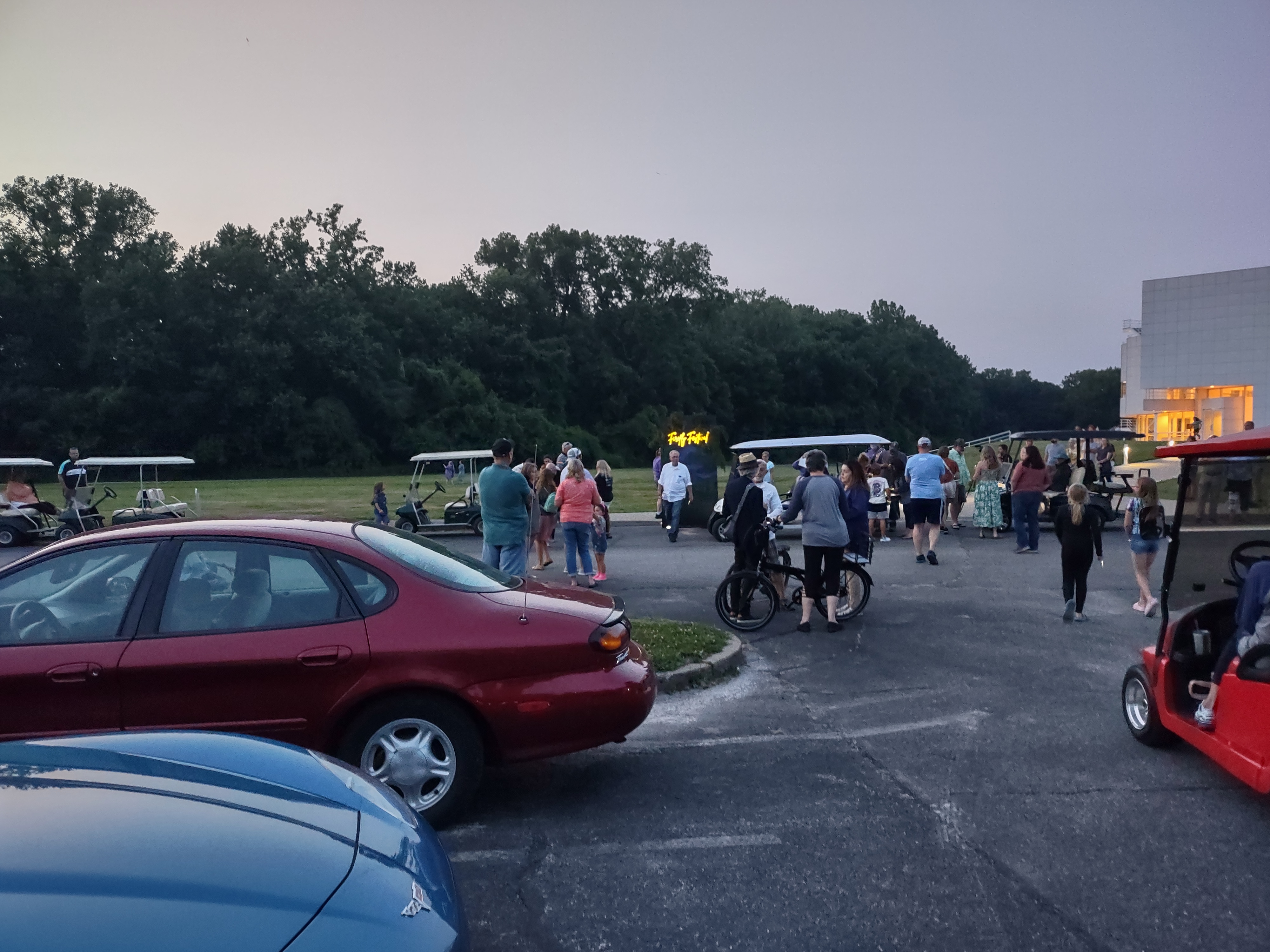 Visitors walking between parked cars and golf carts near event grounds at dusk.