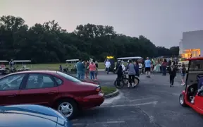 Visitors walking between parked cars and golf carts near event grounds at dusk.