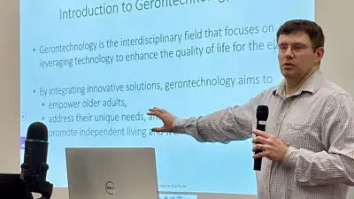 Dr. Ryan Integlia in front of a screen that reads, "Intro to Gerontechnology: Gerontechnology is the interdisciplinary field that focuses on leveraging technology to enhance the quality of life for the elderly. By integrating innovative solutions gerontology aims to empower older adults, address their unique needs, and promote independent living" 