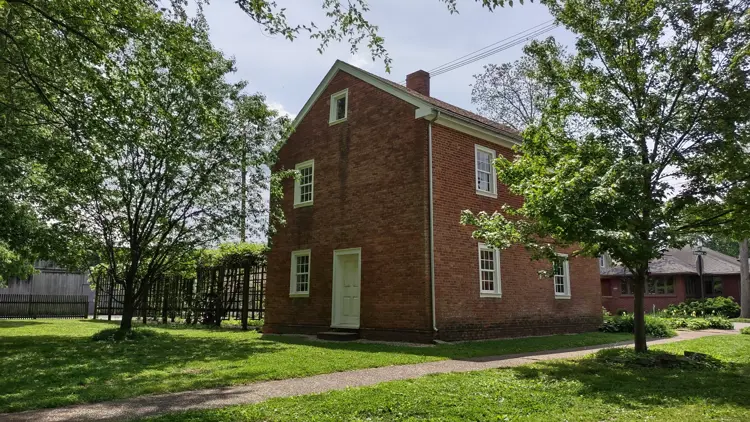 A two-story historic red brick house with white trim stands among trees, with a trellis structure and walkway visible beside the building in a shaded garden setting.