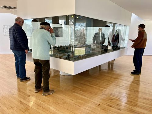 Four visitors stand inside a bright, modern gallery with wood floors, examining a large, glass display case that contains a detailed miniature model of a town. Three visitors on the left look closely at the model, while one visitor on the right gestures toward the display. The glass case reflects the visitors and windows behind them, with trees visible outside.