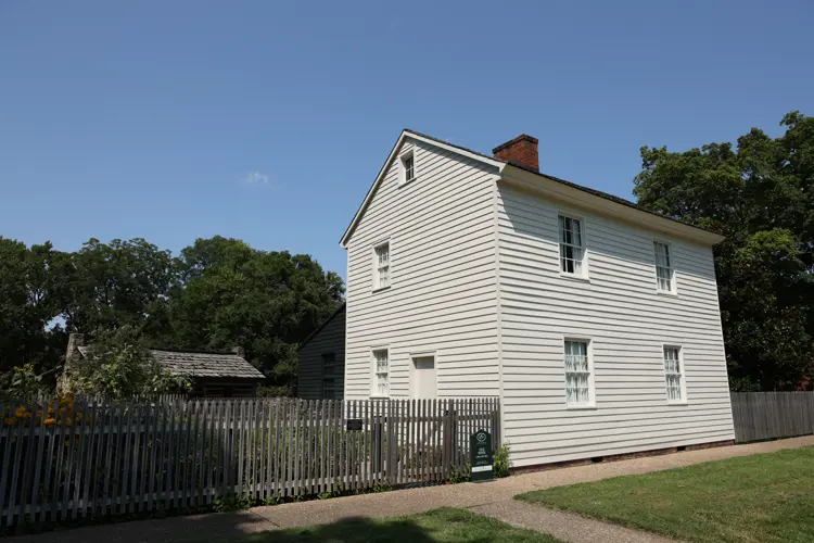 The Lenz House, a white two-story historic building with a pitched roof and chimney, stands behind a wooden picket fence, with a garden and smaller outbuilding visible to the side.