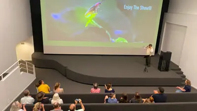 Audience seated in a small theater watching a presentation projected on a large screen showing a firefly and the text “Say’s Firefly, Pyractomena angulata. Enjoy the show!!!” A presenter stands at the front near a podium while attendees sit in tiered seating facing the screen.