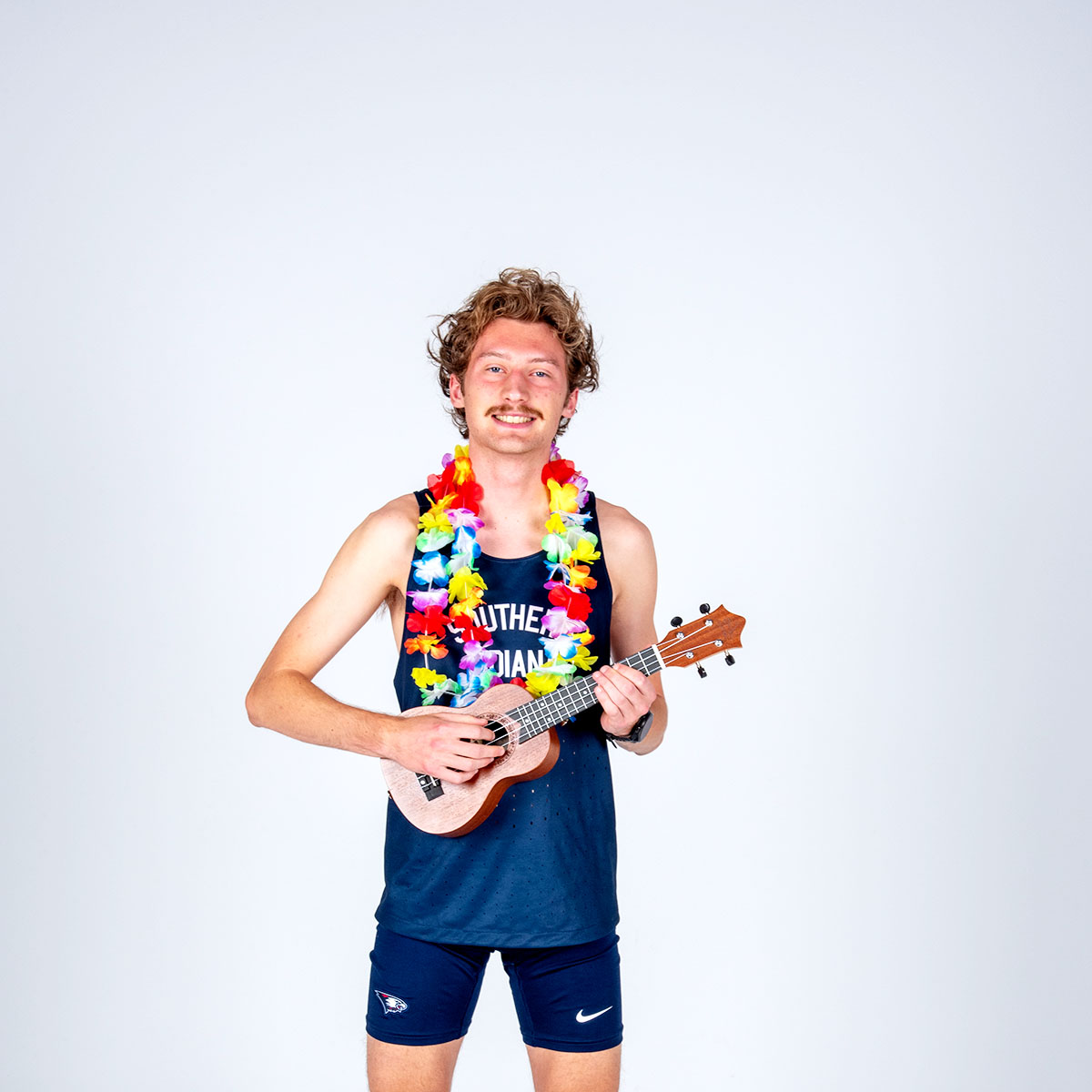A smiling cross country runner wears two colorful leis and strums a ukulele for his media day photo.