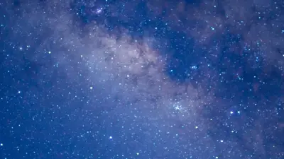 Wide view of a star-filled night sky with the Milky Way visible as a dense, cloudy band of stars stretching across the image in shades of blue and purple.