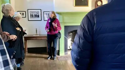 Inside a historic room with wood floors and a green fireplace, a guide stands near the hearth addressing a small group of visitors gathered along the walls. Framed photos and a portrait hang above the mantel, and a wooden table beneath displays additional framed images. The visitors face the guide, listening during the interior portion of a tour.