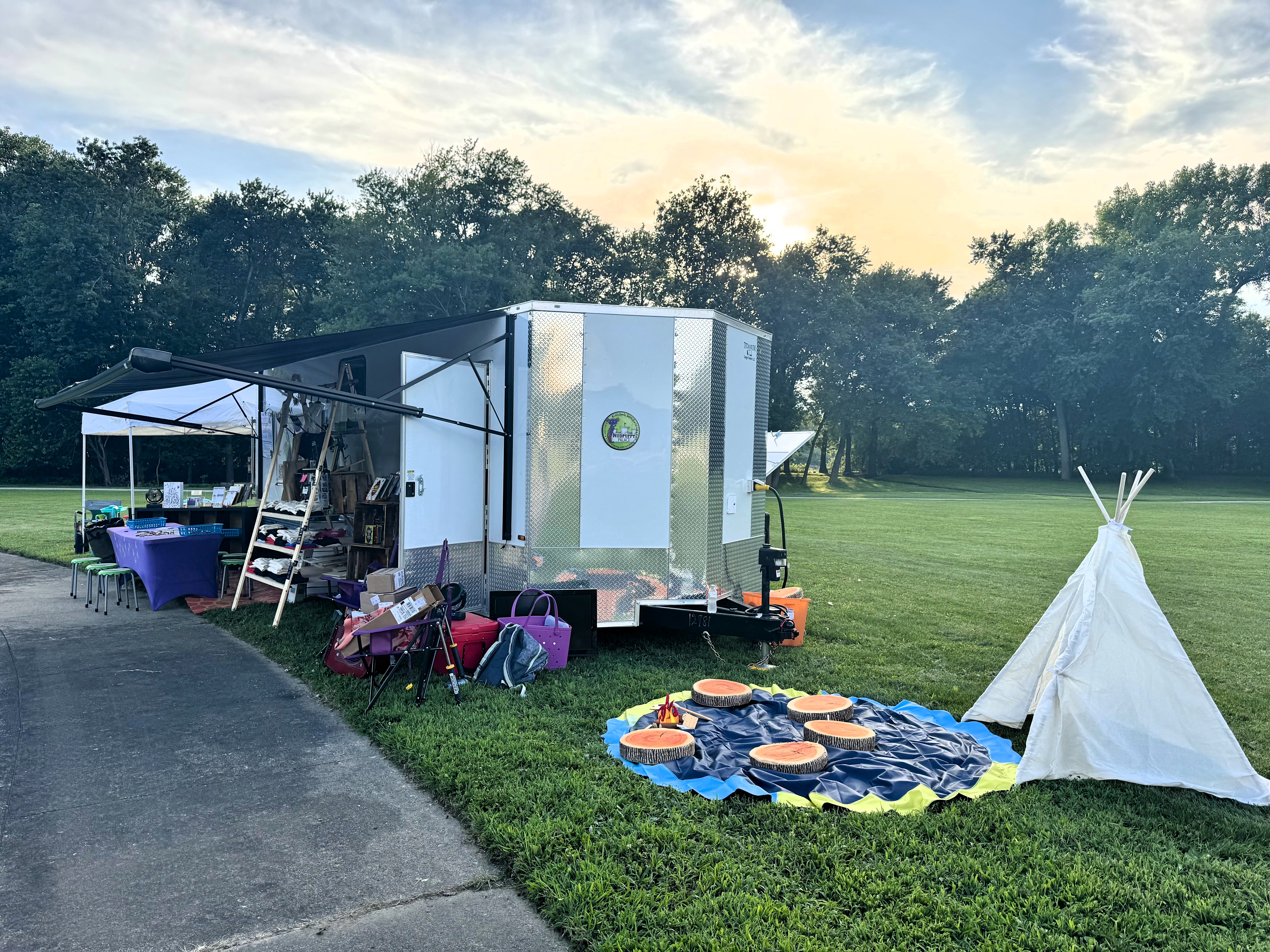 Outdoor activity area with a small white teepee, colorful stepping pads, and a trailer setup on grass.