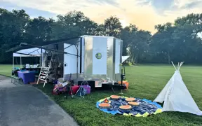 Outdoor activity area with a small white teepee, colorful stepping pads, and a trailer setup on grass.