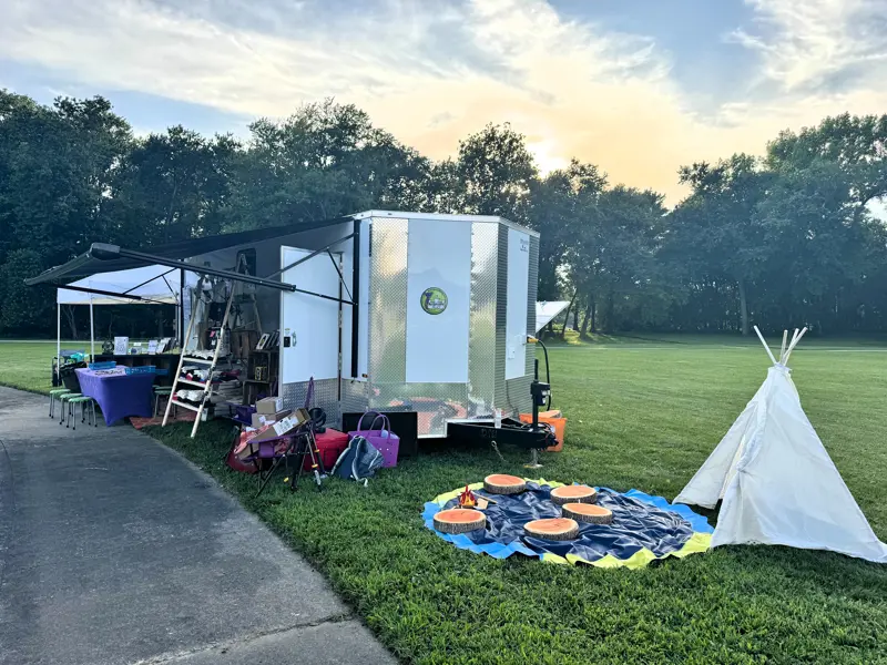 Outdoor activity area with a small white teepee, colorful stepping pads, and a trailer setup on grass.