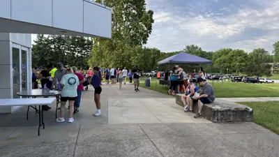 Visitors gathered outside the Atheneum Visitor Center in New Harmony during the Firefly Festival, with people standing near tables, sitting on a low concrete wall, and walking along a paved path beside a grassy lawn and canopy tent.