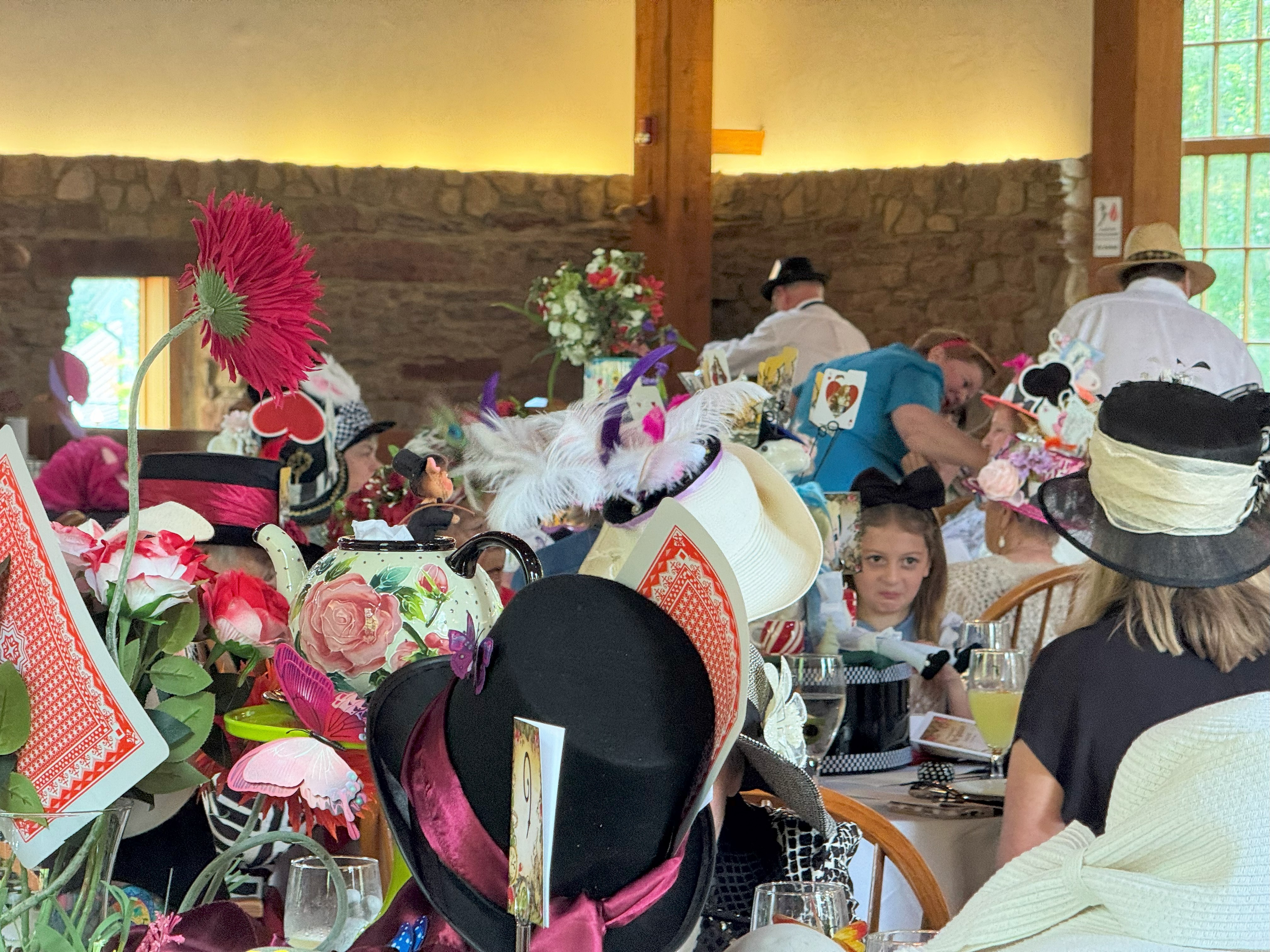 Attendees sit at decorated tables during a themed luncheon, wearing elaborate hats with playing card and floral designs, in a rustic room with stone walls and large windows.