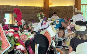 Attendees sit at decorated tables during a themed luncheon, wearing elaborate hats with playing card and floral designs, in a rustic room with stone walls and large windows.