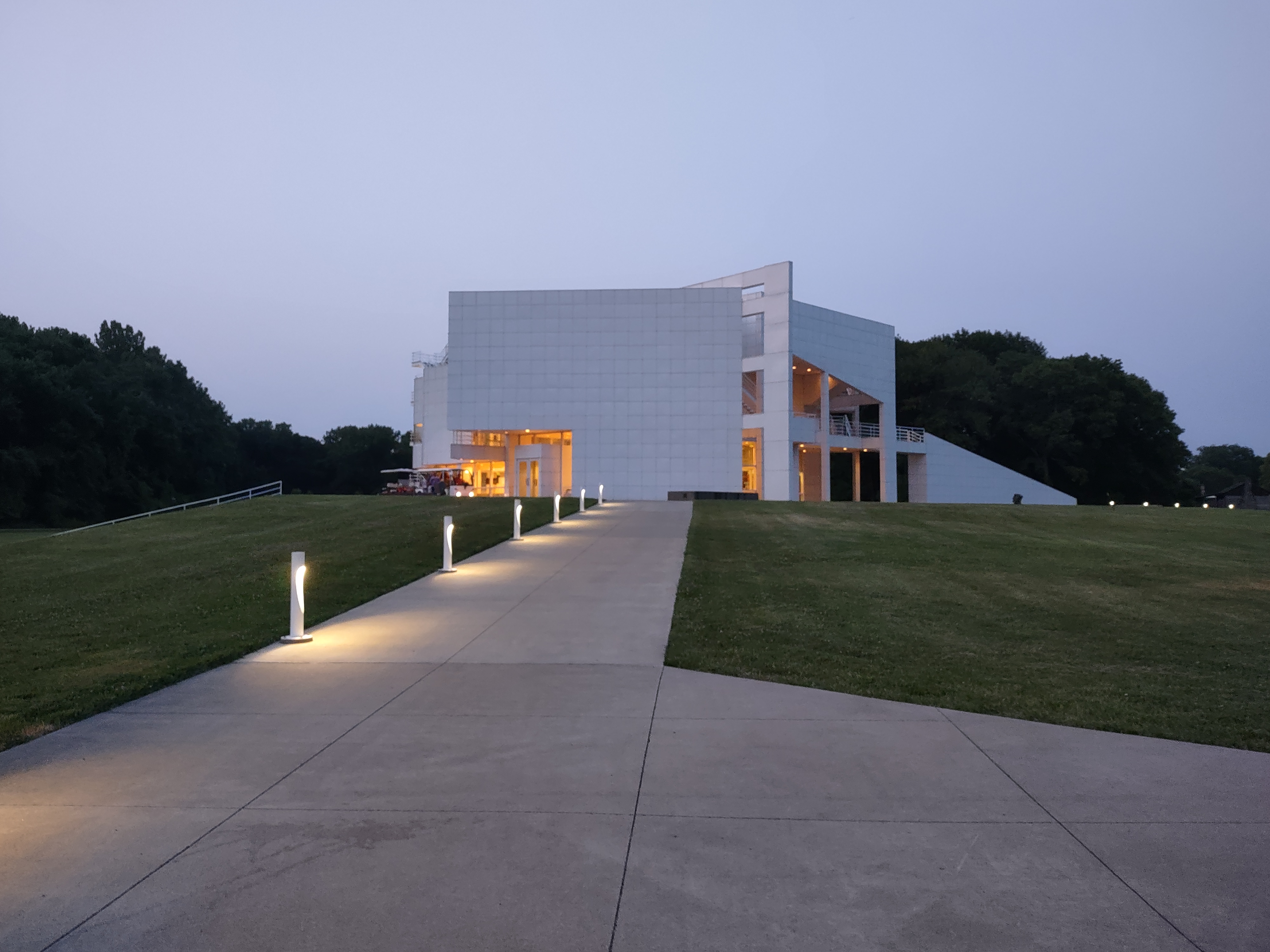 Modern white building lit from inside at dusk with a walkway leading up to it.