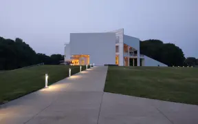Modern white building lit from inside at dusk with a walkway leading up to it.