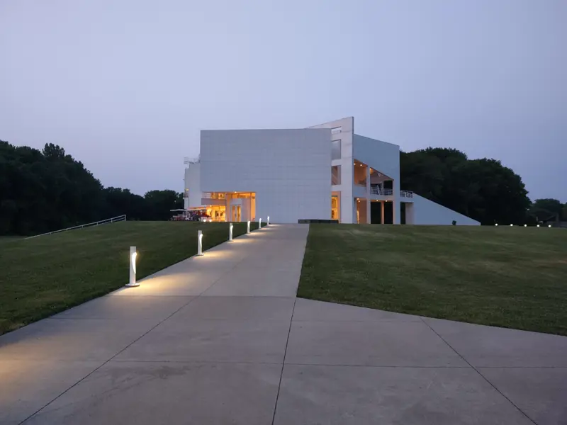 Modern white building lit from inside at dusk with a walkway leading up to it.
