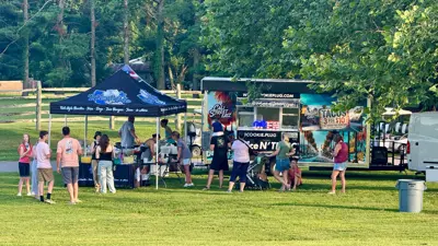 Outdoor festival scene with a food truck and a pop-up tent set up on a grassy lawn. Several visitors are gathered nearby—some standing in line at the truck, others seated on the grass—while trees and greenery surround the area in the background.