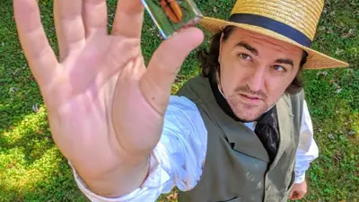 Person standing outdoors on grass, holding up a small clear case containing an orange-and-black insect toward the camera. The person wears a straw hat, vest, and boots, and is positioned below the camera angle, with trees and greenery in the background.