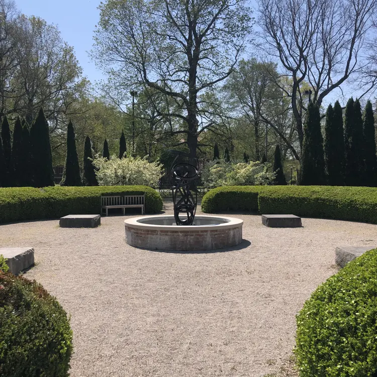 A circular garden setting features a central abstract metal sculpture rising from a low brick basin, surrounded by gravel paths, trimmed hedges, benches, and tall trees in the background under a clear sky.
