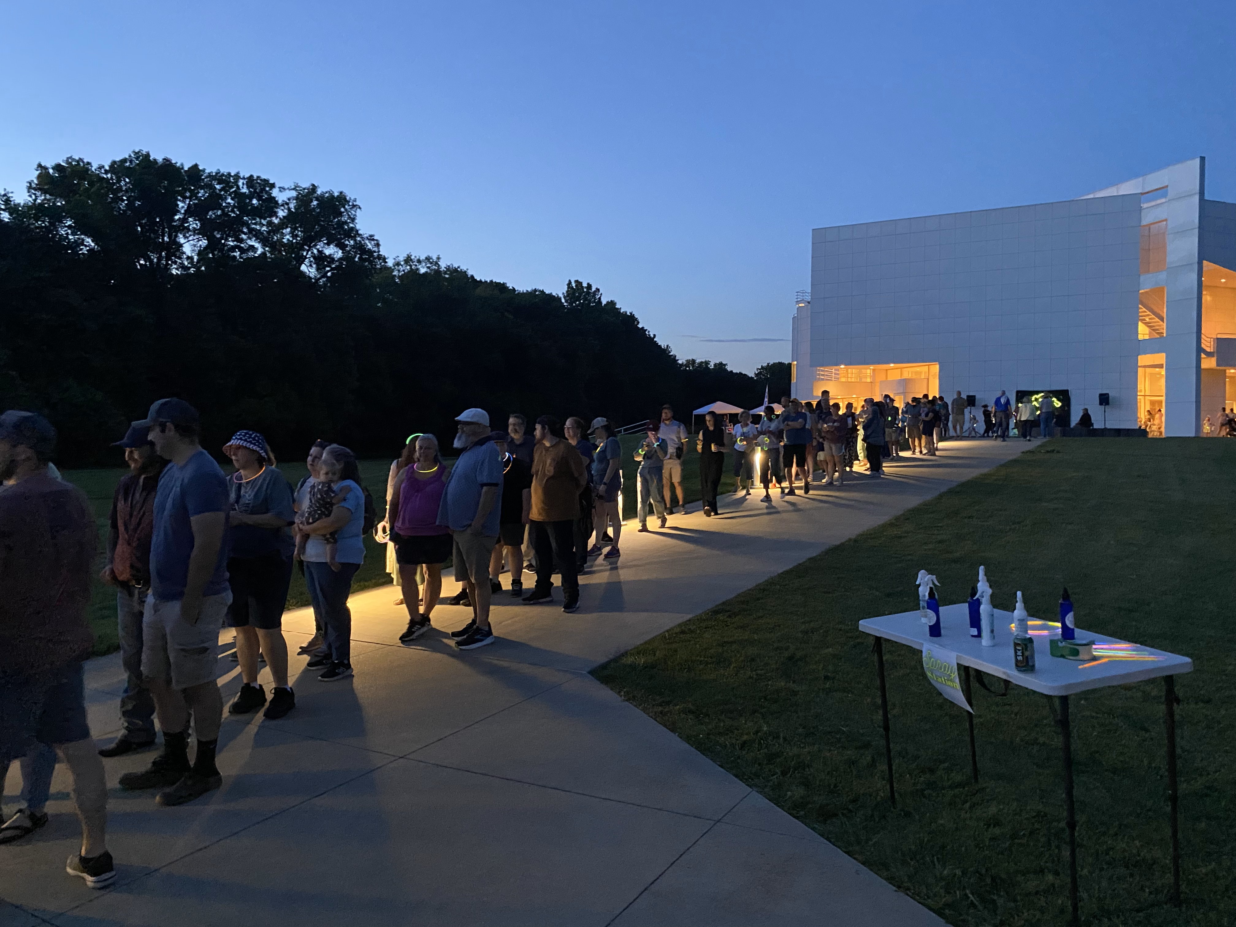 Line of people outside a modern building at dusk with illuminated interior visible.