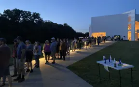 Line of people outside a modern building at dusk with illuminated interior visible.