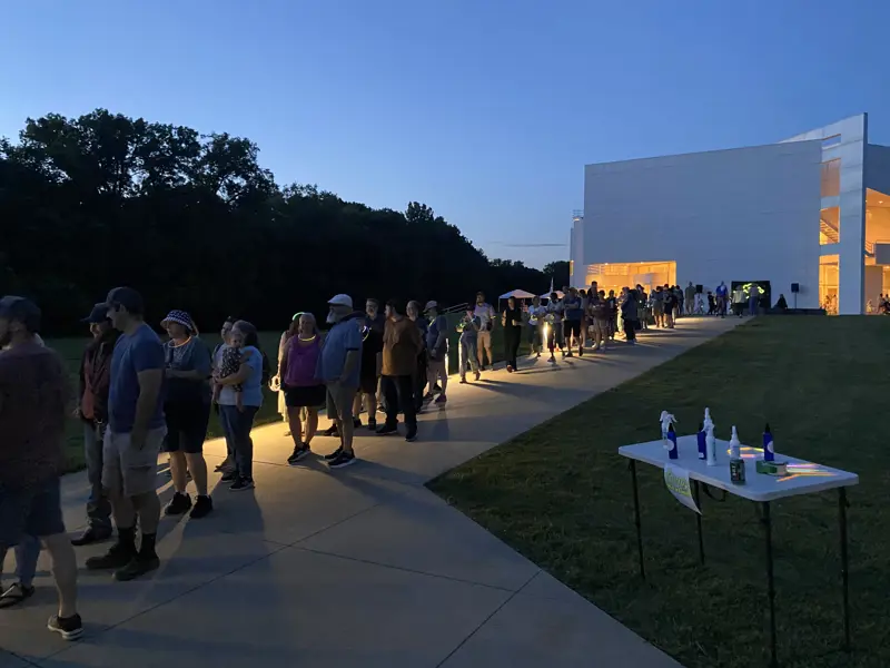Line of people outside a modern building at dusk with illuminated interior visible.