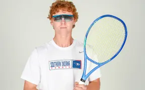 A member of the tennis team, wearing sunglasses, poses with his racquet, in the studio during tennis media day.