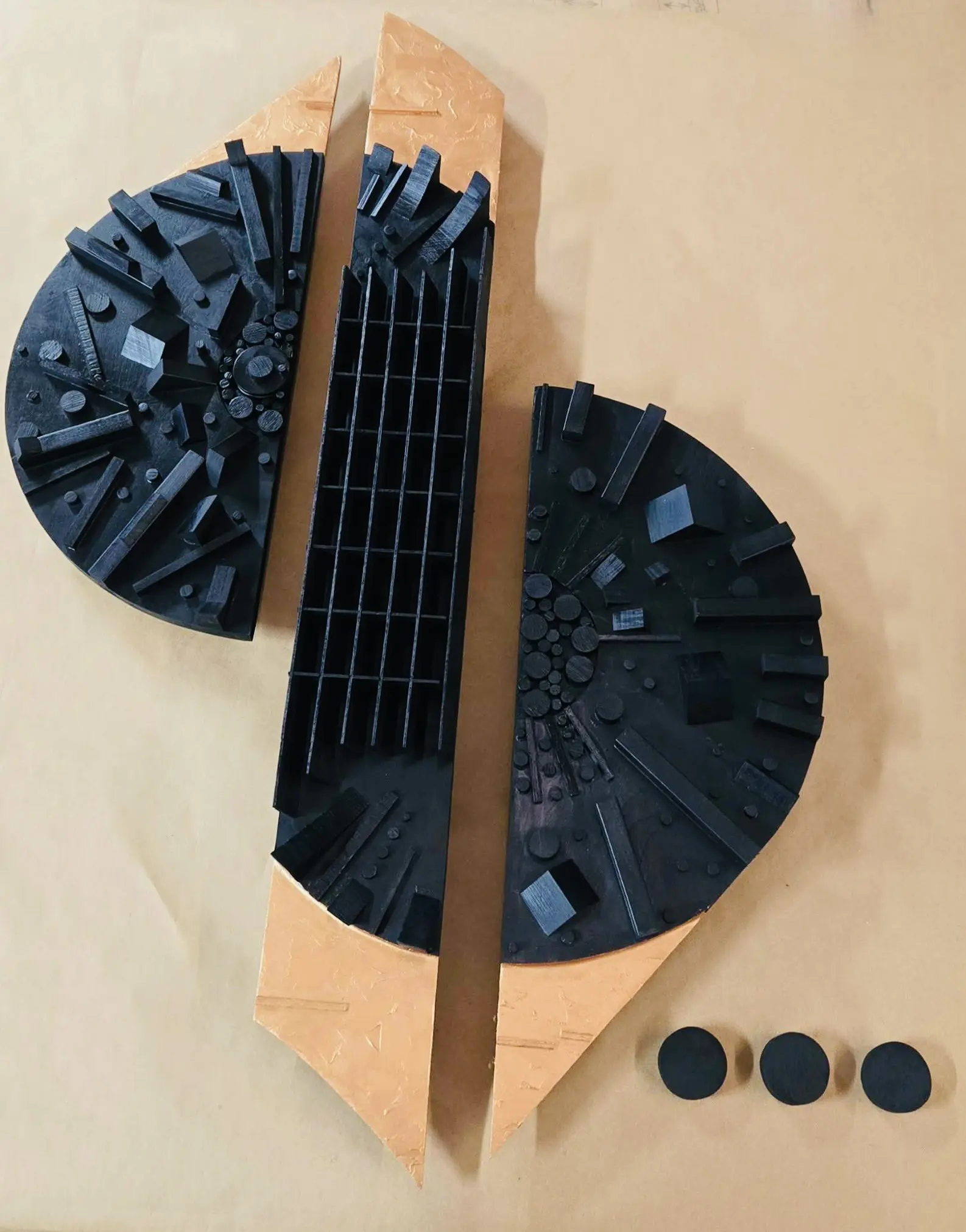 The image shows the work Inner City (2026) by Amy Musia. It is Reclaimed wood scrap 48 x 32 x 4” arranged in varying heights close together - as if a city -- making two half circles together painted black and disconnected by another black strip of irregular depth going vertical. Triangles of flat wood painted bronze are attached alternating the bottom and top of the half circles and rectangle in the middle, as if empty lots or room to expand.