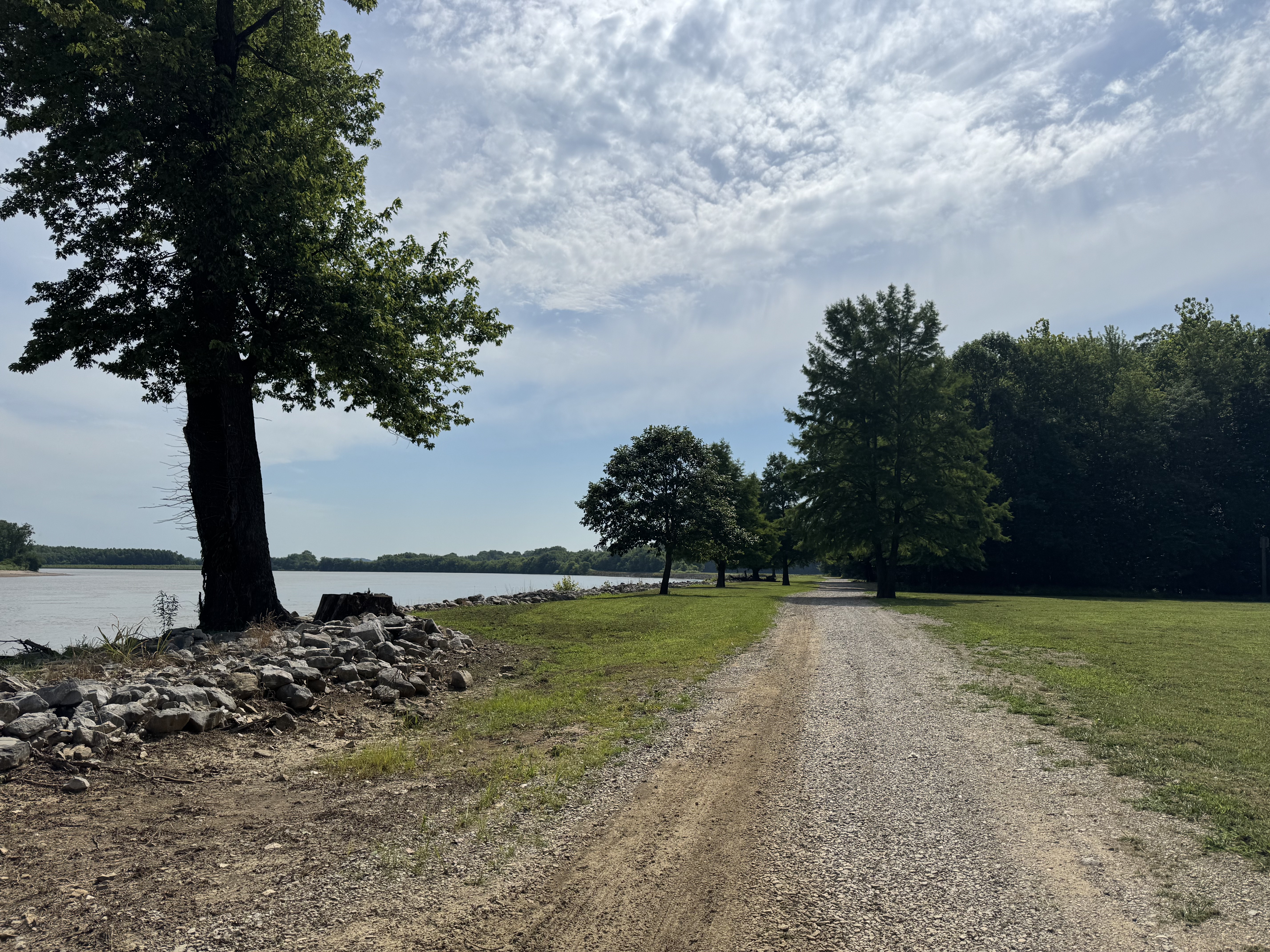 A gravel path runs alongside a wide river, bordered by grass and trees, with a line of rocks along the shoreline and a partly cloudy sky stretching overhead.