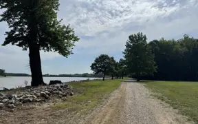 A gravel path runs alongside a wide river, bordered by grass and trees, with a line of rocks along the shoreline and a partly cloudy sky stretching overhead.