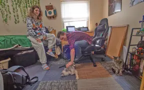 A student leans over to pet his friend's small dog in his campus room, while his cat looks on. 