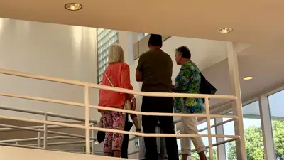 Four visitors stand on an upper-level indoor balcony with curved white railings, looking out over a bright, modern space with large windows. One person stands near the center facing a display or point of interest, while the others gather close by, listening. Below, additional visitors can be seen near the entrance, and outside the windows are trees and a grassy area under clear daylight.